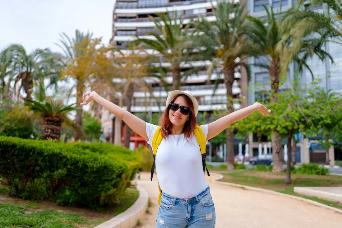 Happy solo female traveler posing with open arms in a palm-lined area of Alicante.