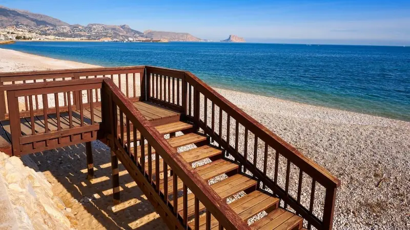 Wooden stairs leading down to Playa Cap Blanc in El Campello, Alicante, with clear Mediterranean waters and a quiet beach atmosphere