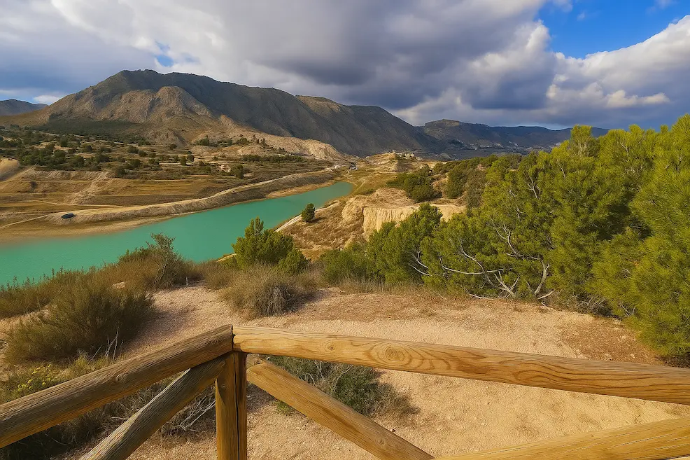 Panoramic view of the turquoise river and mountain landscape near Sella, Alicante. The image shows a wooden viewpoint overlooking pine trees, rugged hills, and the Sierra de Aitana under a partly cloudy sky. A scenic spot in the interior of Costa Blanca ideal for hiking and nature photography.